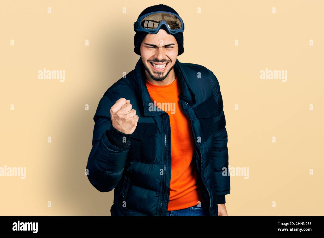 Young hispanic man with beard wearing snow wear and sky glasses angry and mad raising fist frustrated and furious while shouting with anger. rage and Stock Photo