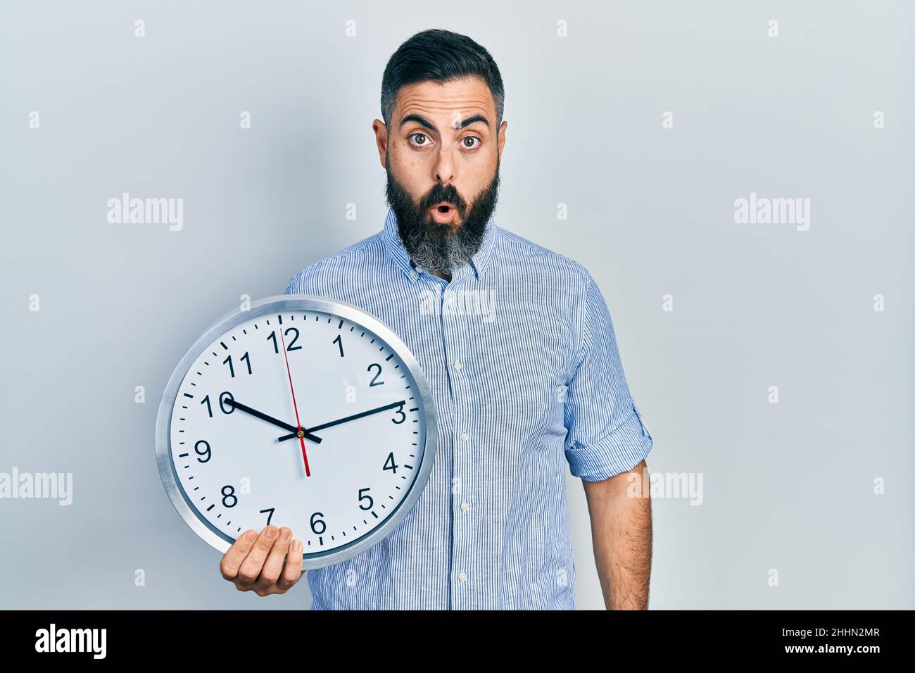 Young hispanic man holding big clock scared and amazed with open mouth