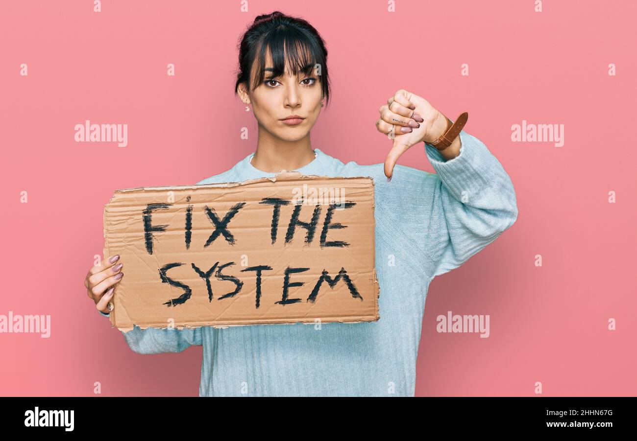 Young hispanic woman holding fix the system banner cardboard with angry face, negative sign showing dislike with thumbs down, rejection concept Stock Photo