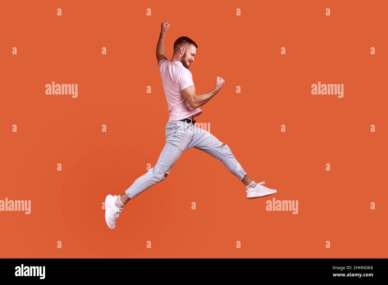 Side view portrait of happy bearded man jumping in air showing yes i did it gesture, copy space for ad, wearing pink T-shirt and jeans. Indoor studio shot isolated on orange background. Stock Photo