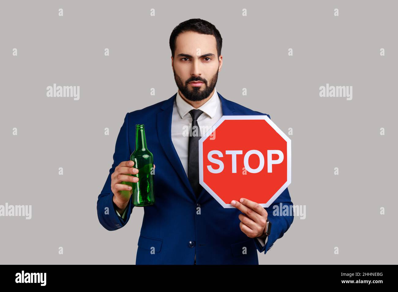 Portrait of serious strict bearded man showing alcoholic beverage beer bottle and stop sign, warning and worrying, wearing official style suit. Indoor studio shot isolated on gray background. Stock Photo