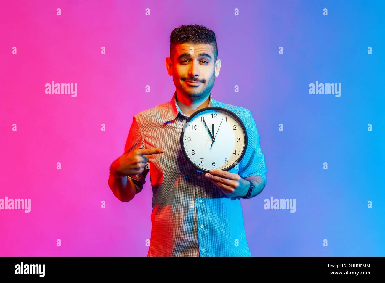 Look at time. Portrait of handsome positive man in shirt pointing big clock and looking at camera, showing wall watches to hurry up. Indoor studio shot isolated on colorful neon light background. Stock Photo