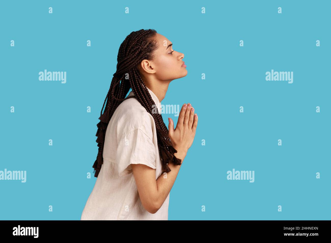 Side view of woman with dreadlocks keeps palms pressed together, pleading angel, has innocent expressions, asks for apologize, wearing white shirt. Indoor studio shot isolated on blue background. Stock Photo