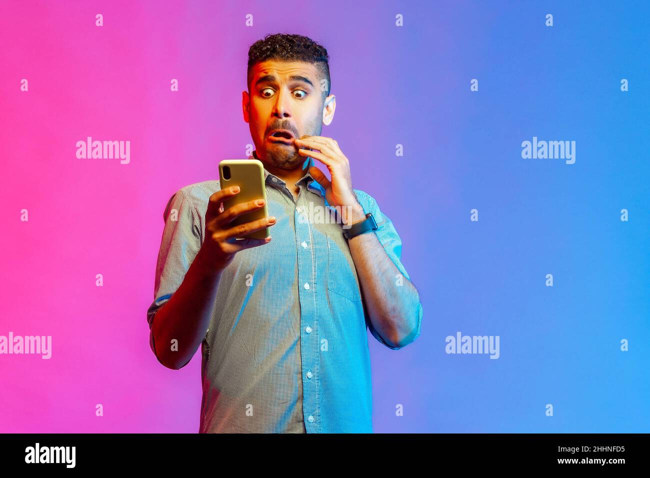 Portrait of man in shirt looking at smartphone display with funny scared face, shocked and surprised with application. Indoor studio shot isolated on colorful neon light background. Stock Photo