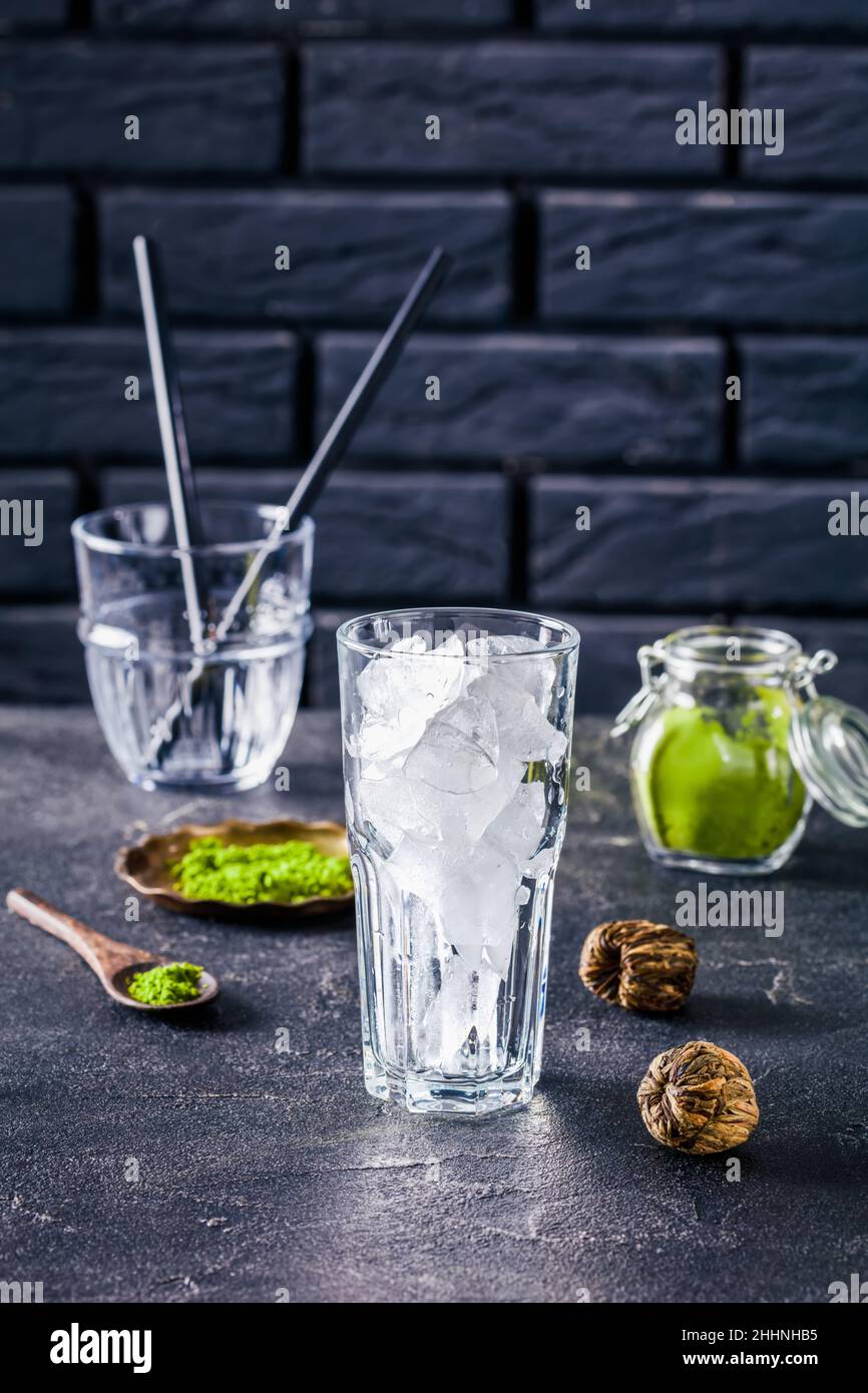 a glass with ice cubes with a brick wall and powdered matcha tea in glass jar at the background, close up, vertical view Stock Photo
