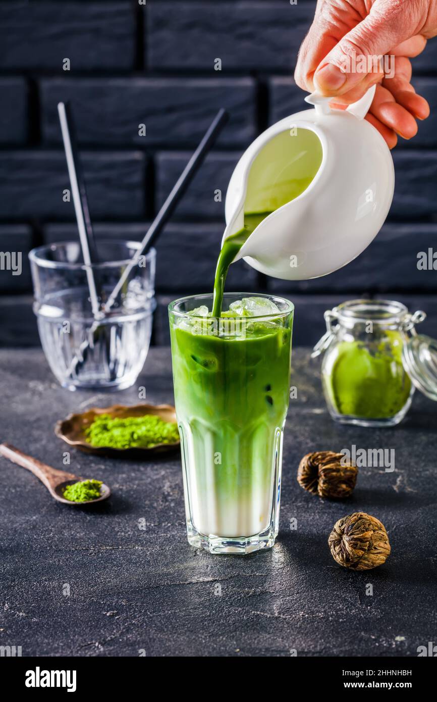 barista hand pouring matcha tea in a glass with matcha latte mixed with ice cubes with a brick wall at the background and powdered matcha tea in glass Stock Photo