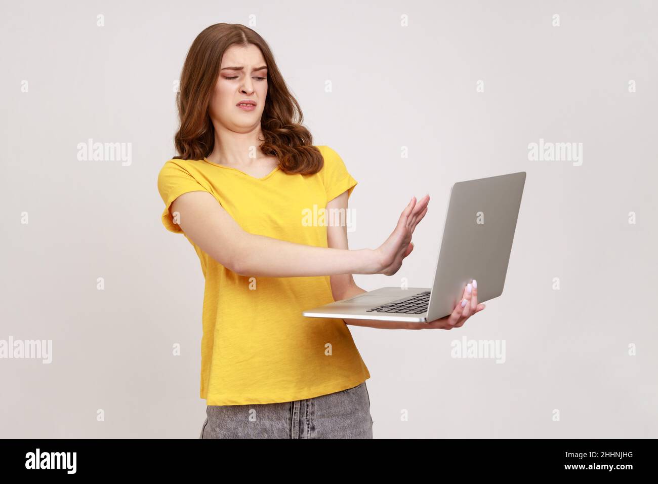 Forbidden content. Frightened teenager girl closing laptop and keeping hand in stop gesture to cover shameful post on internet, troubles with computer. Indoor studio shot isolated on gray background. Stock Photo