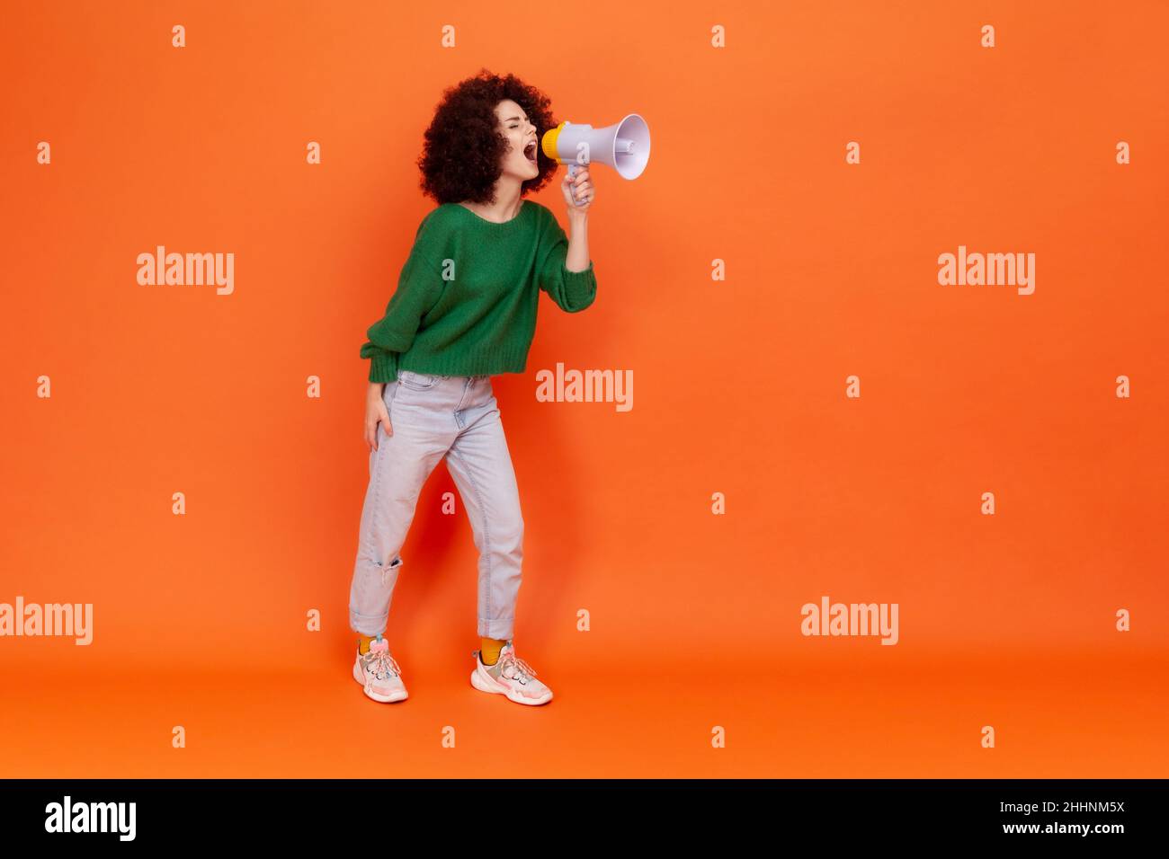 Full length portrait of woman with Afro hairstyle wearing green casual style sweater standing screaming loud in megaphone, protesting. Indoor studio shot isolated on orange background. Stock Photo