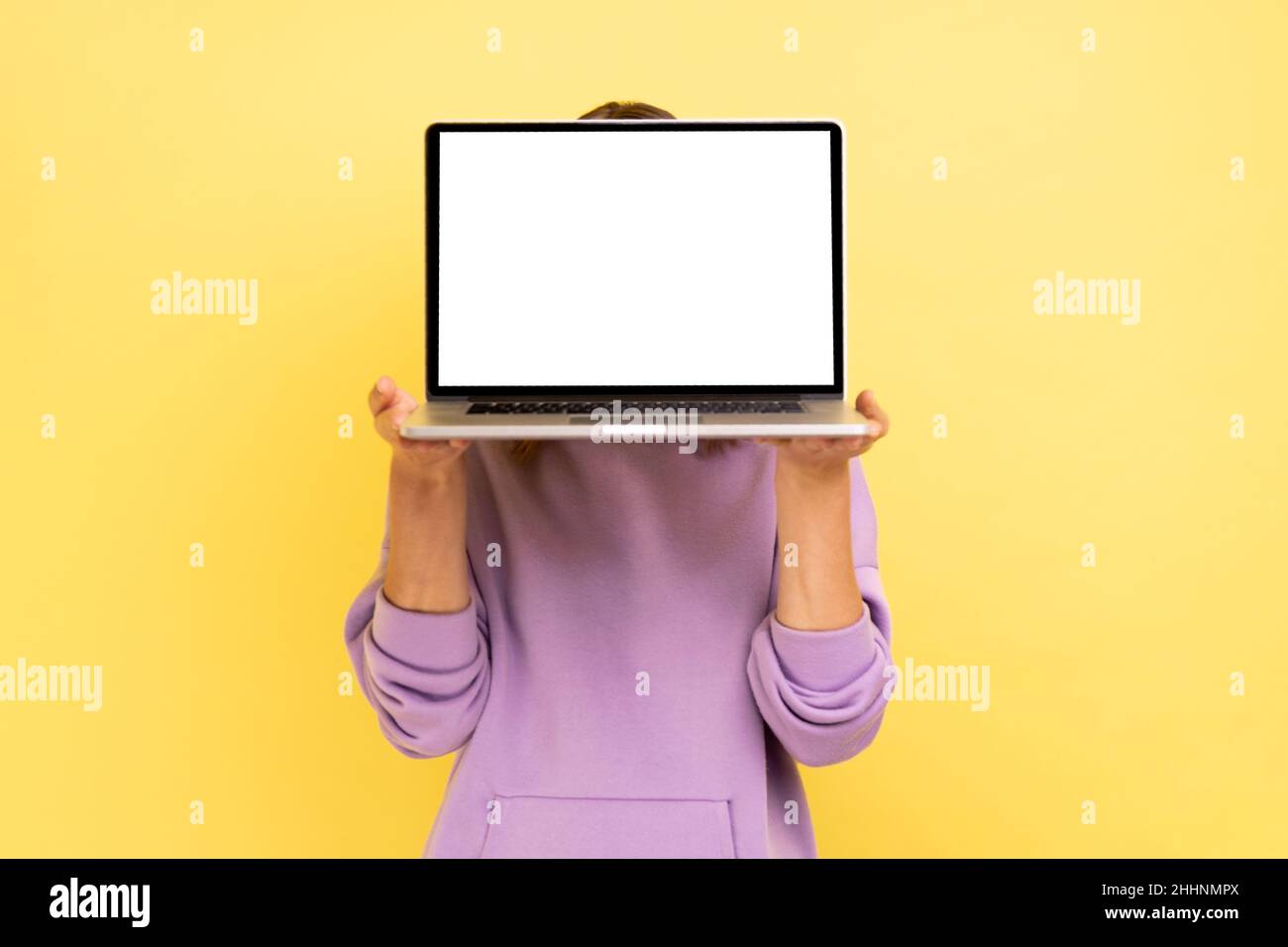 Portrait of unrecognizable woman hiding her face behind laptop with blank display for advertisement or promotional text, wearing purple hoodie. Indoor studio shot isolated on yellow background. Stock Photo