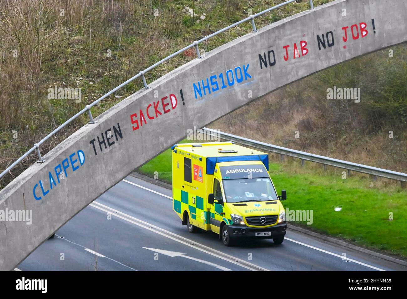 Weymouth, Dorset, UK.  25th January 2022.  An NHS Ambulance passes under a bridge on the A354 at Weymouth in Dorset which has had a graffiti message written on it saying “Clapped Then Sacked! NHS100K No Jabs No Jobs!  The message has appeared as the deadline for the governments mandatory Covid vaccination for NHS staff approaches with the deadline for the first jab on 3rd February and for them to be fully vaccinated by 1st April 2022.  Picture Credit: Graham Hunt/Alamy Live News Stock Photo