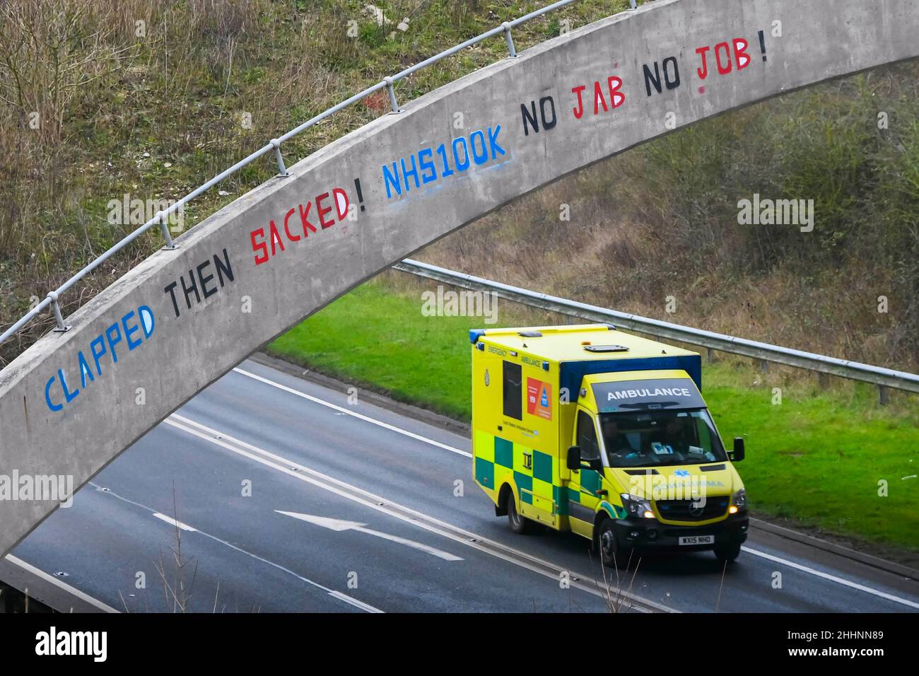 Weymouth, Dorset, UK.  25th January 2022.  An NHS Ambulance passes under a bridge on the A354 at Weymouth in Dorset which has had a graffiti message written on it saying “Clapped Then Sacked! NHS100K No Jabs No Jobs!  The message has appeared as the deadline for the governments mandatory Covid vaccination for NHS staff approaches with the deadline for the first jab on 3rd February and for them to be fully vaccinated by 1st April 2022.  Picture Credit: Graham Hunt/Alamy Live News Stock Photo