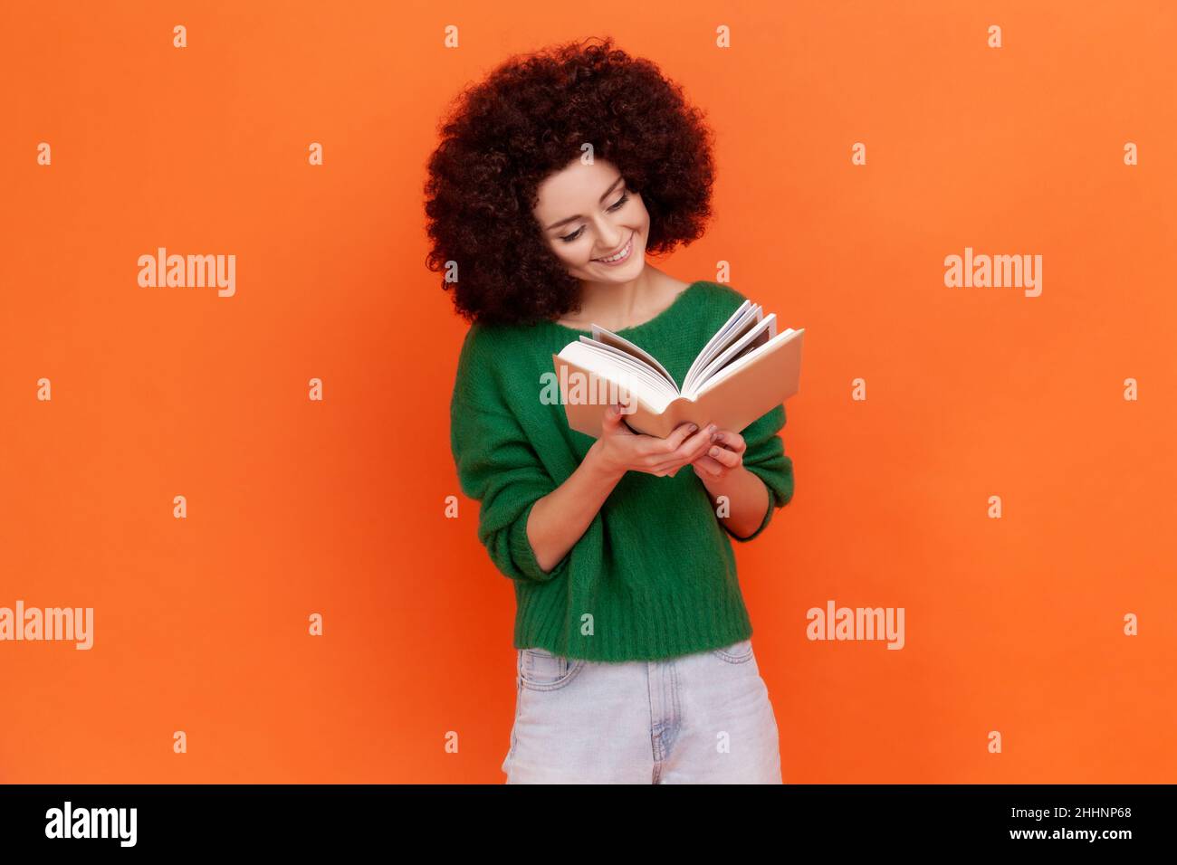 Happy smiling woman with Afro hairstyle wearing green casual style sweater reading book with interesting plot, studying with pleasure. Indoor studio shot isolated on orange background. Stock Photo