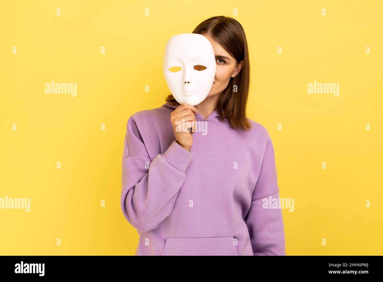 Woman with dark hair removing white mask from face showing his smiling expression, good mood, pretending to be another person, wearing purple hoodie. Indoor studio shot isolated on yellow background. Stock Photo