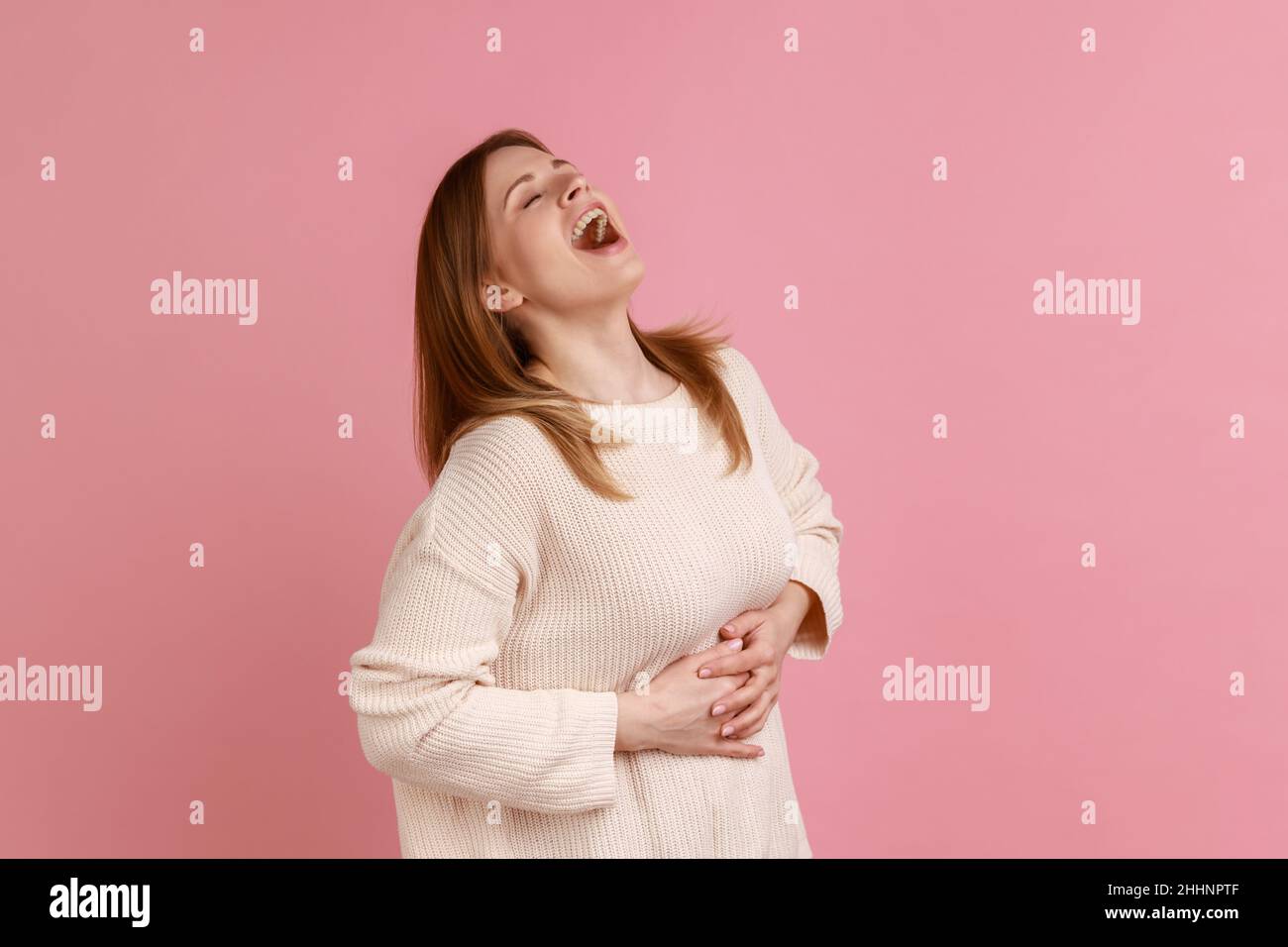 Portrait of happy blond woman holding belly, laughing out loud, taunting with hysterical laughter, mocking friend, wearing white sweater. Indoor studio shot isolated on pink background. Stock Photo