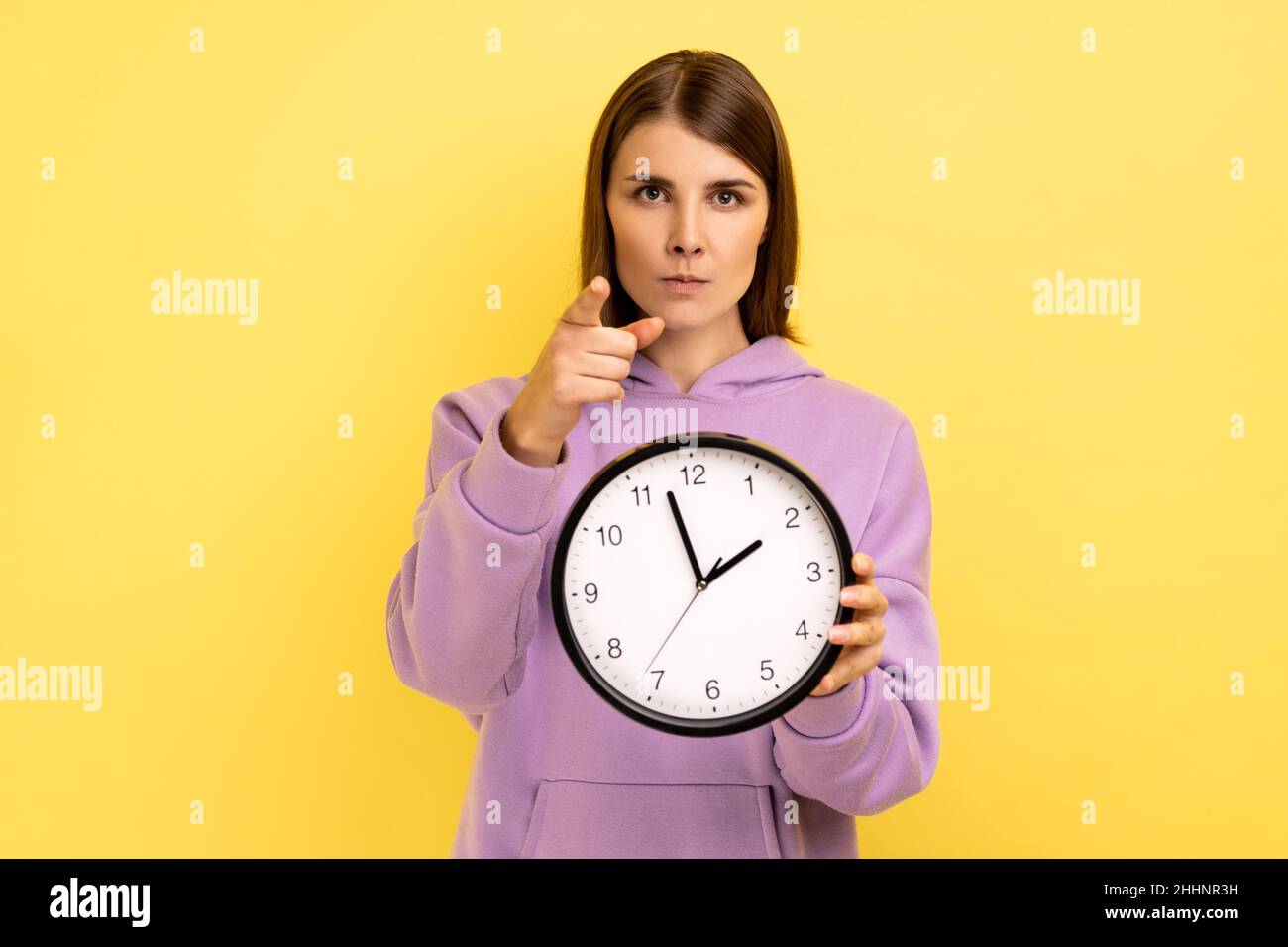 Strict dark haired young adult woman holding wall clock in hands and pointing to camera at you, time management, you are late, wearing purple hoodie. Indoor studio shot isolated on yellow background. Stock Photo