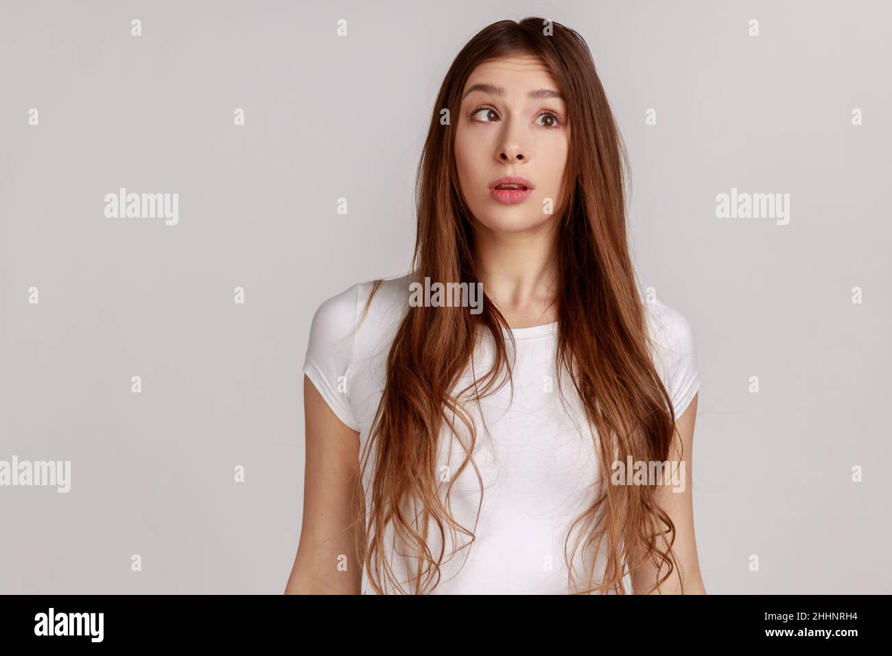 Funny young woman with dark hair making silly comical face with eyes crossed, thinking intensely looking dumb and confused, wearing white T-shirt. Indoor studio shot isolated on gray background. Stock Photo