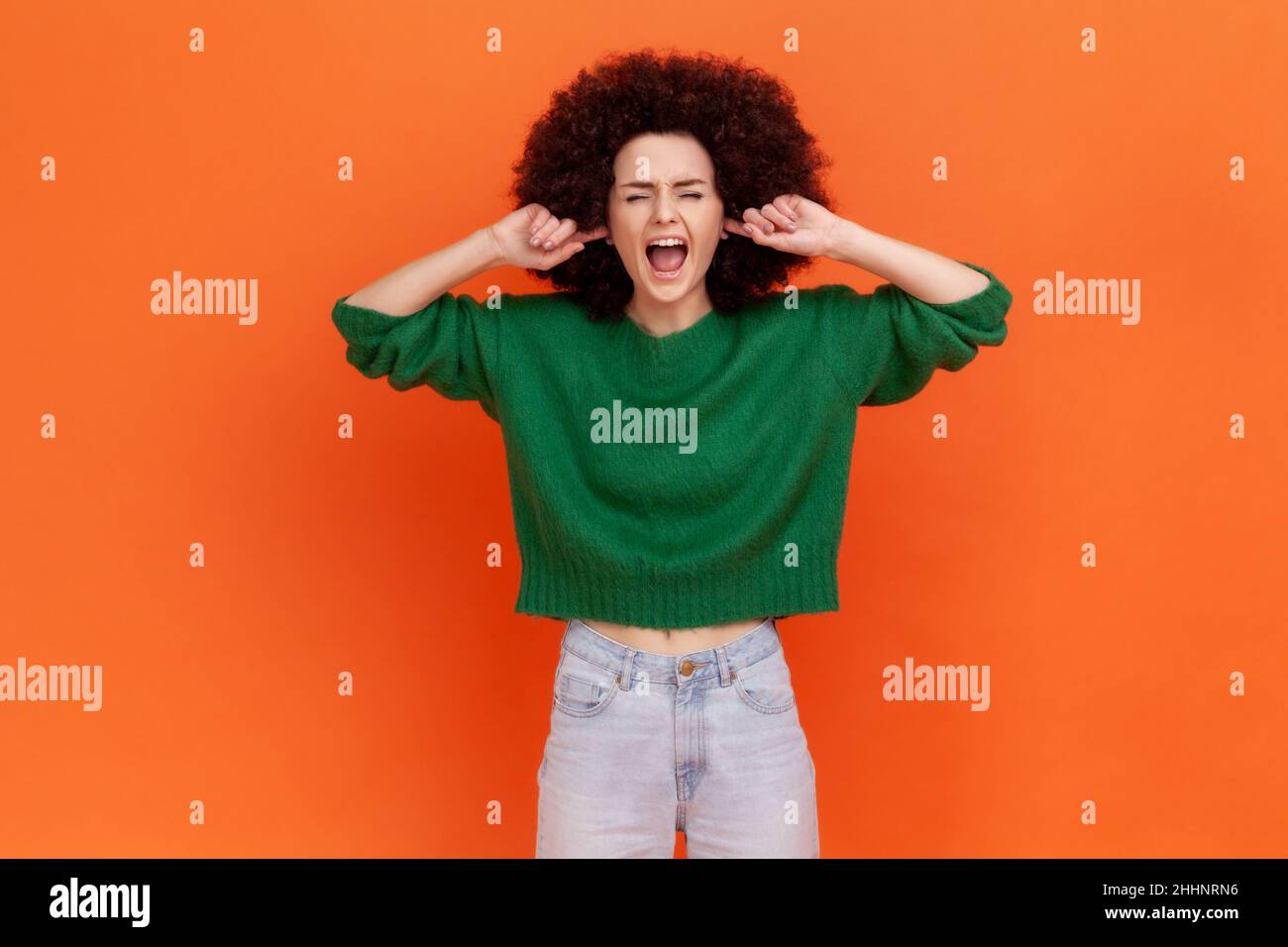 Irritated woman with Afro hairstyle wearing green sweater closing her eyes and ears with fingers, tired of bothersome loud sound, high decibel alarm. Indoor studio shot isolated on orange background. Stock Photo