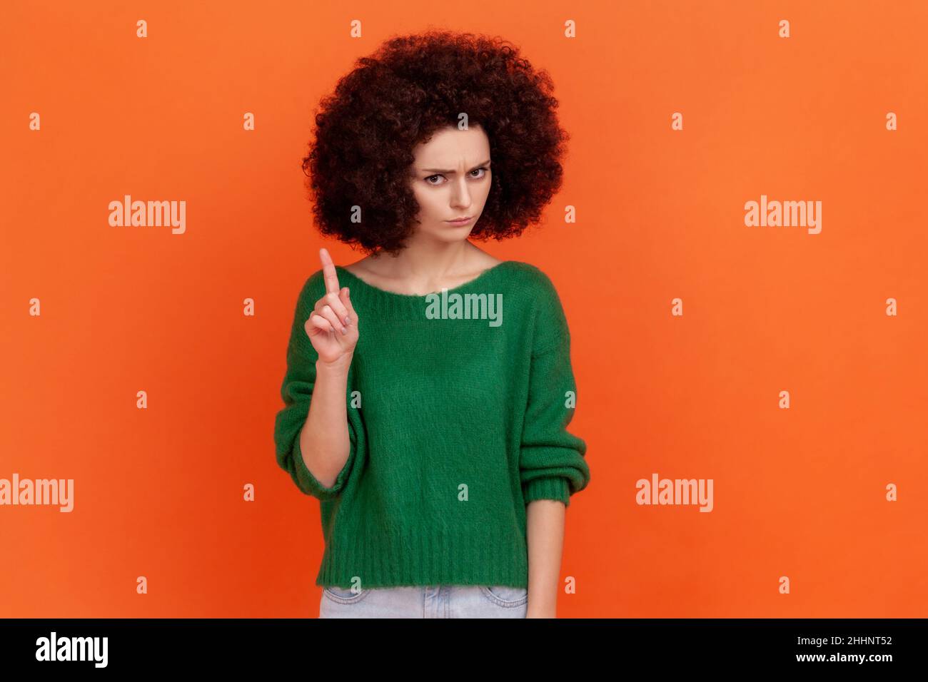 Portrait of serious woman with Afro hairstyle wearing green casual style sweater looking at camera, showing warning attention sign. Indoor studio shot isolated on orange background. Stock Photo