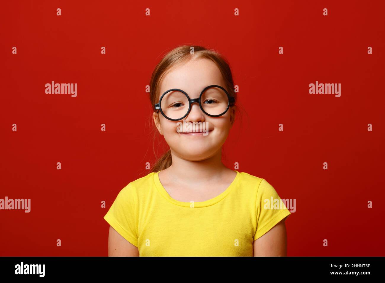 Funny little girl with glasses. Schoolgirl child in a yellow t-shirt on a red isolated background. Stock Photo