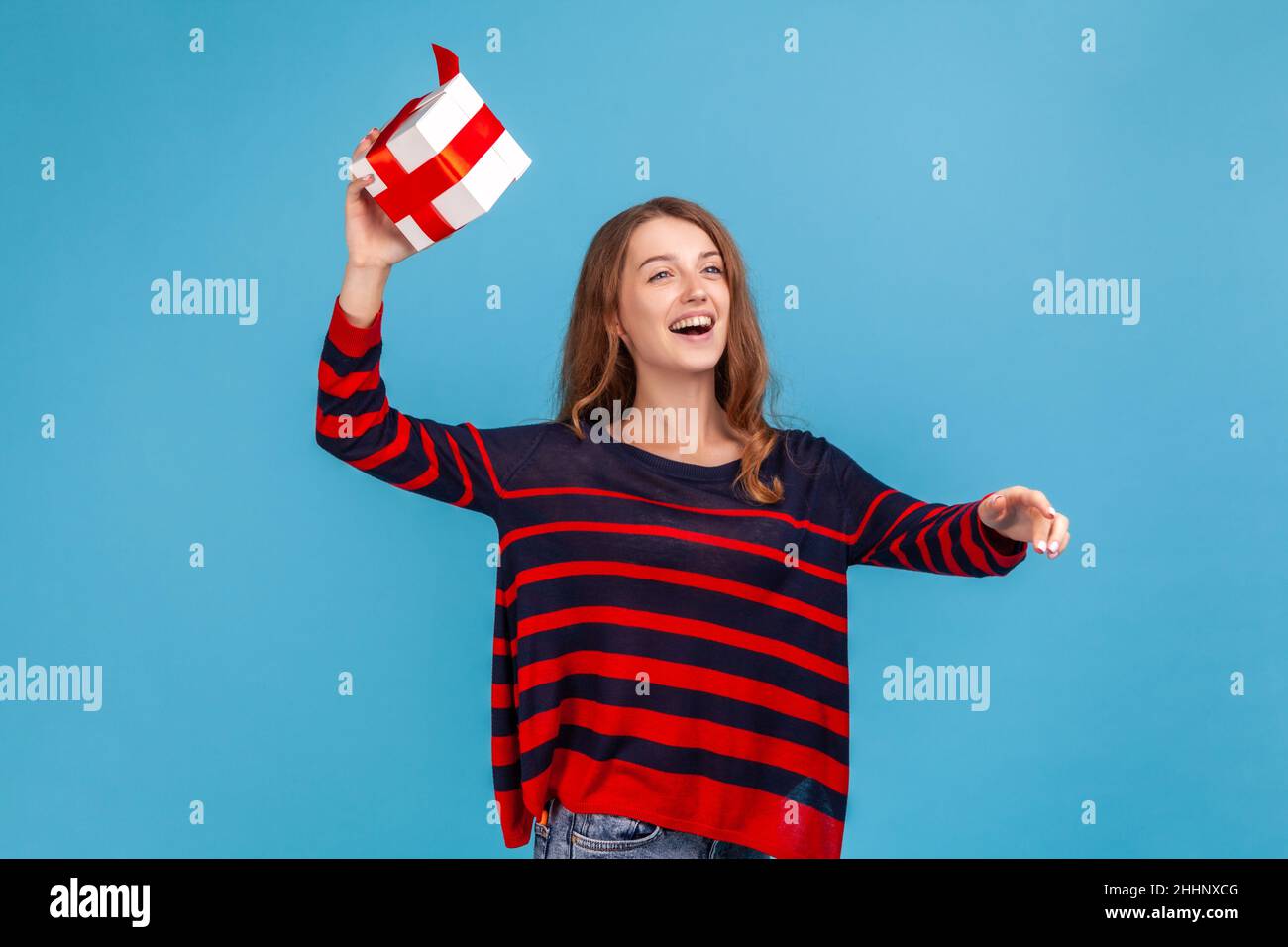 Happy excited woman wearing striped casual style sweater, throwing present box, having happy look, celebrating holiday, congratulating. Indoor studio shot isolated on blue background. Stock Photo