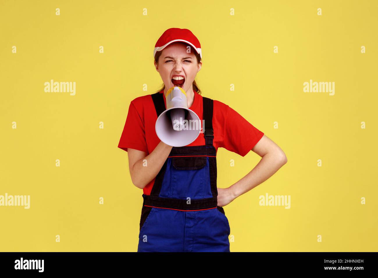 Portrait of angry worker woman screaming something with angry facial expression, protesting, keeps hand on hip, wearing overalls and red cap. Indoor studio shot isolated on yellow background. Stock Photo