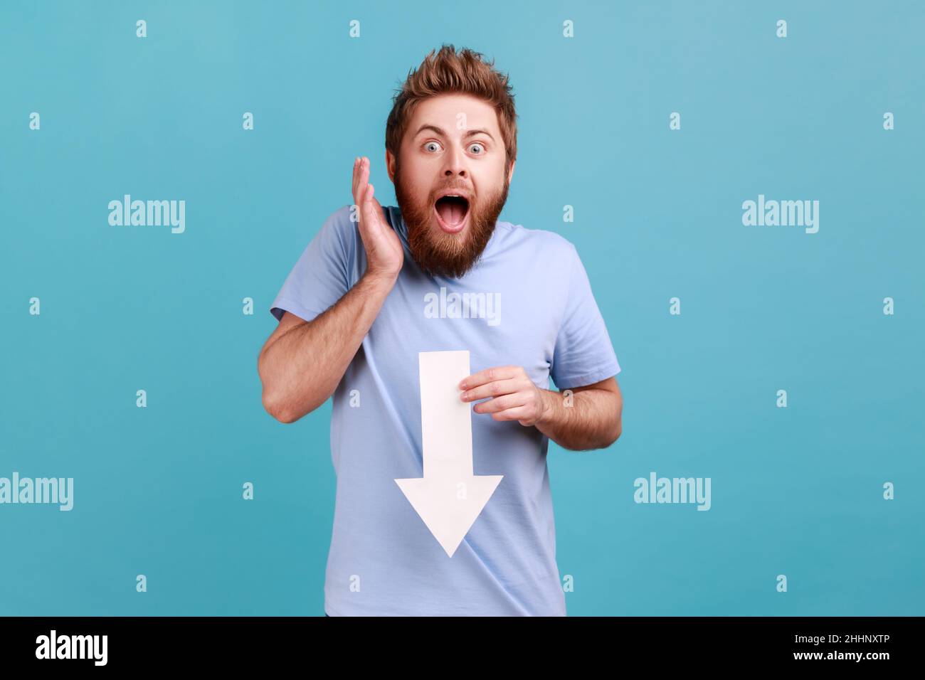 Portrait of astonished surpised bearded man showing white arrow pointing down, expressing shocked emotions, downgrade concept. Indoor studio shot isolated on blue background. Stock Photo