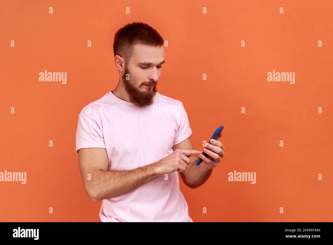 Portrait of bearded man scrolling online, texting message in social media on cell phone, using mobile network services, wearing pink T-shirt. Indoor studio shot isolated on orange background. Stock Photo