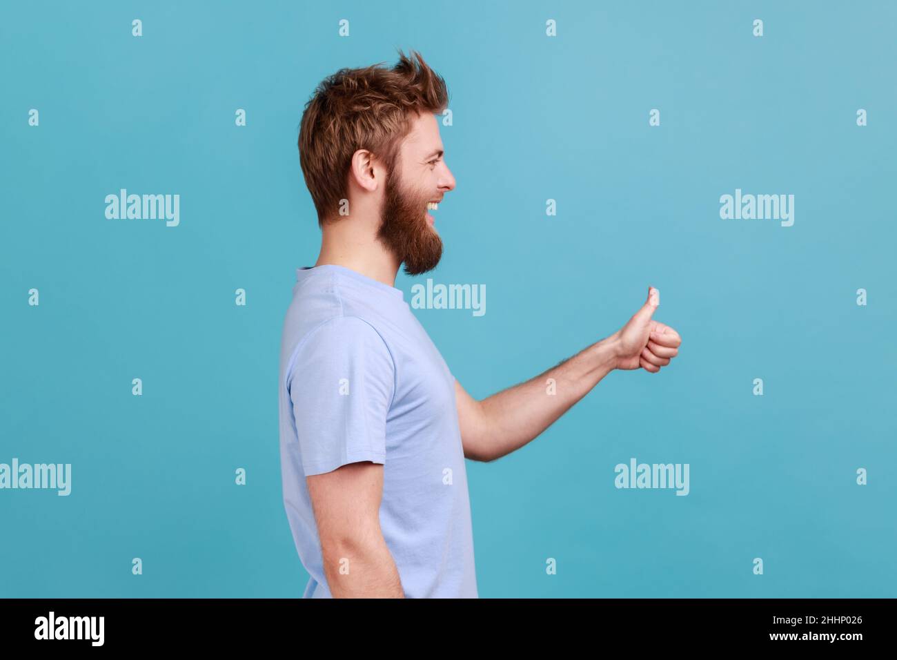 Side view of bearded man approves incredible promo keeps thumbs up likes and agrees being satisfied with something, happy expression. Indoor studio shot isolated on blue background. Stock Photo