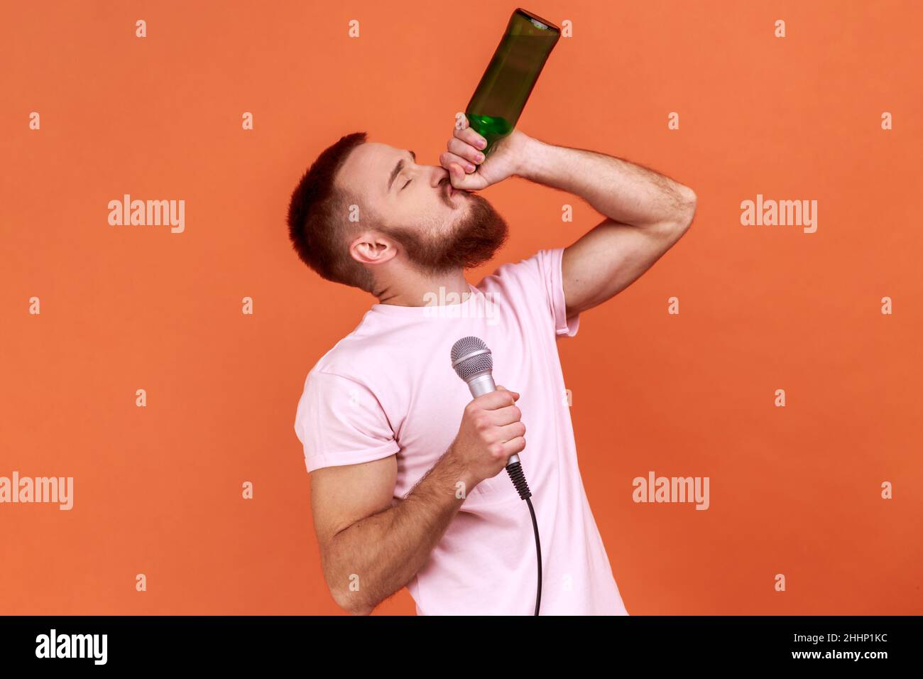 Portrait of drunk bearded man spending time in karaoke, holding microphone in hand, drinking alcoholic beverage, wearing pink T-shirt. Indoor studio shot isolated on orange background. Stock Photo