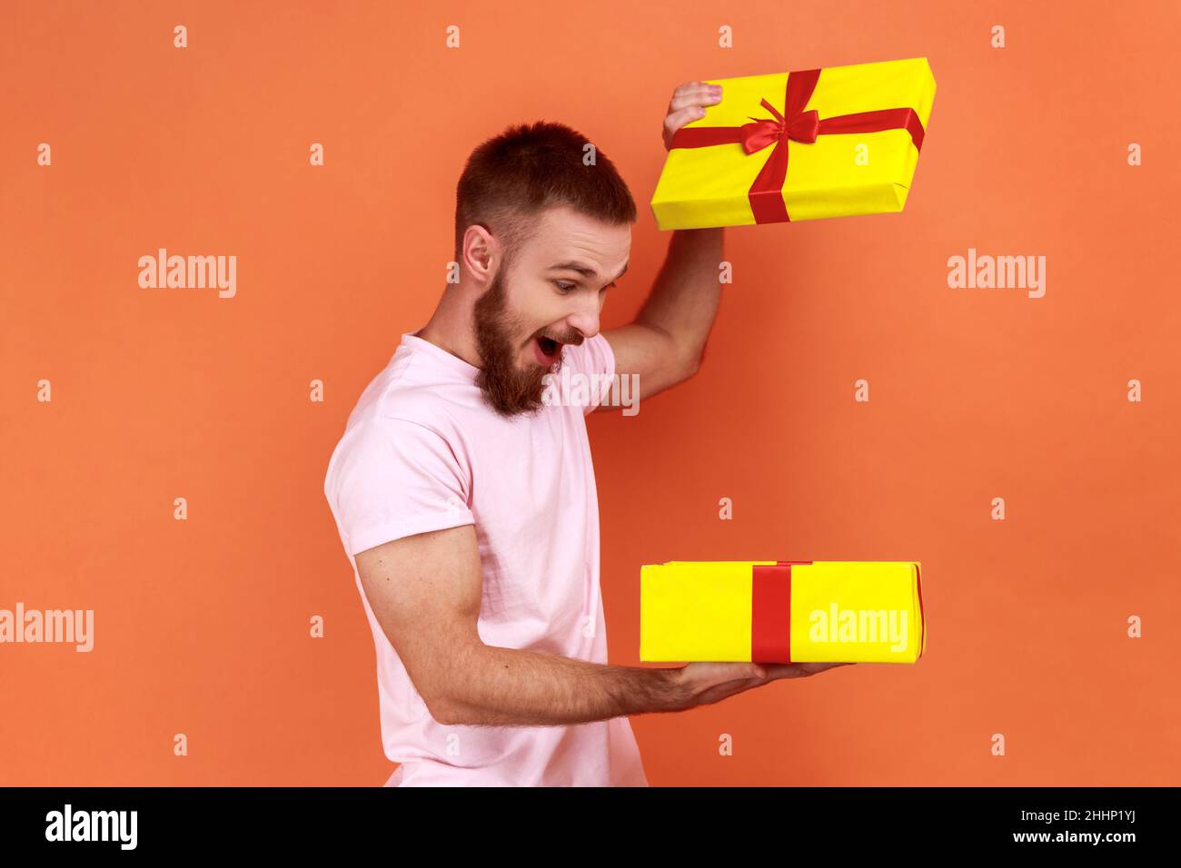 Portrait of handsome bearded man holding in hands unboxed gift, being satisfied with present, being extremely happy, wearing pink T-shirt. Indoor studio shot isolated on orange background. Stock Photo