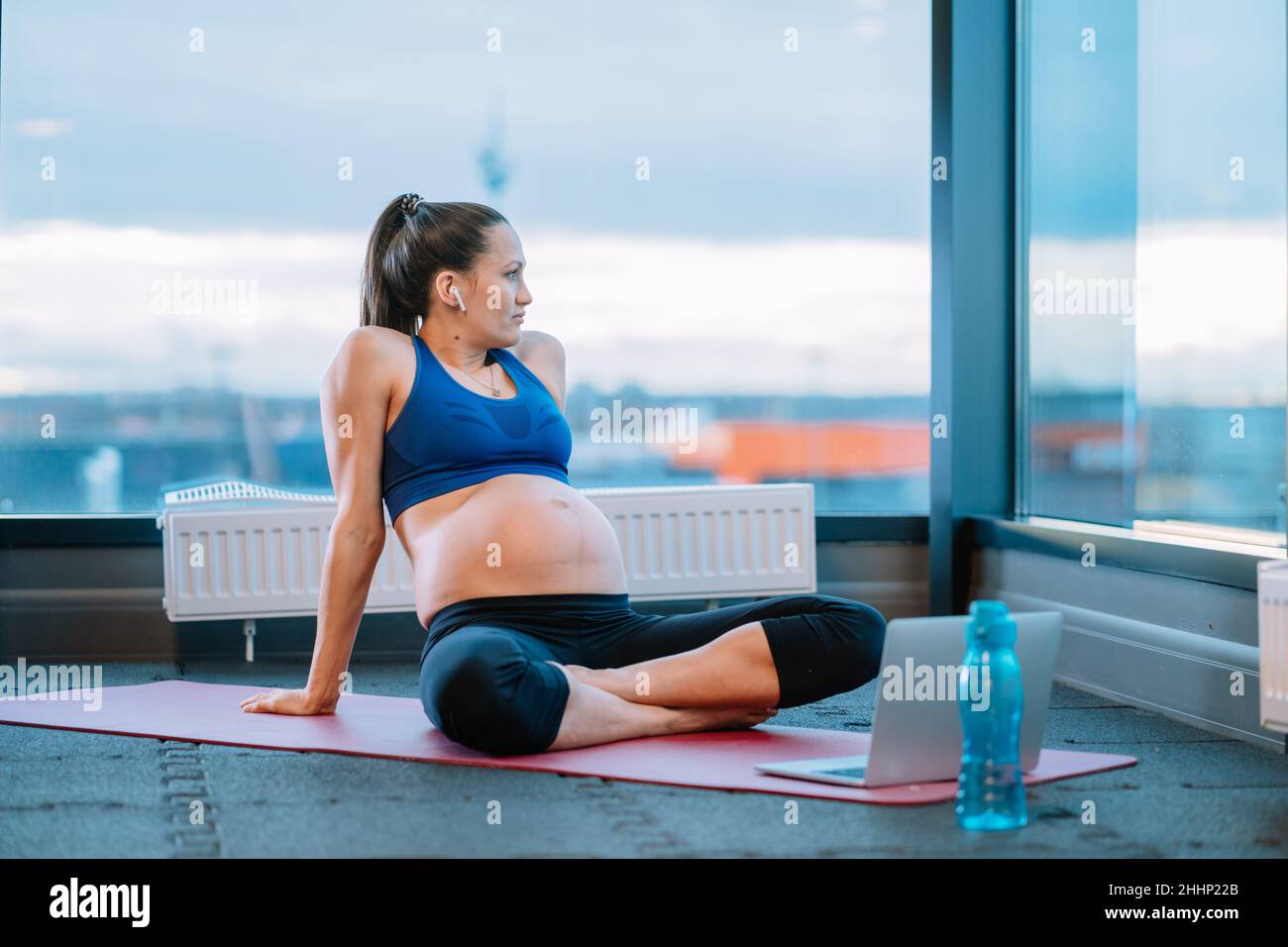 Pregnant woman in wireless earbuds resting during online prenatal workout at gym Stock Photo