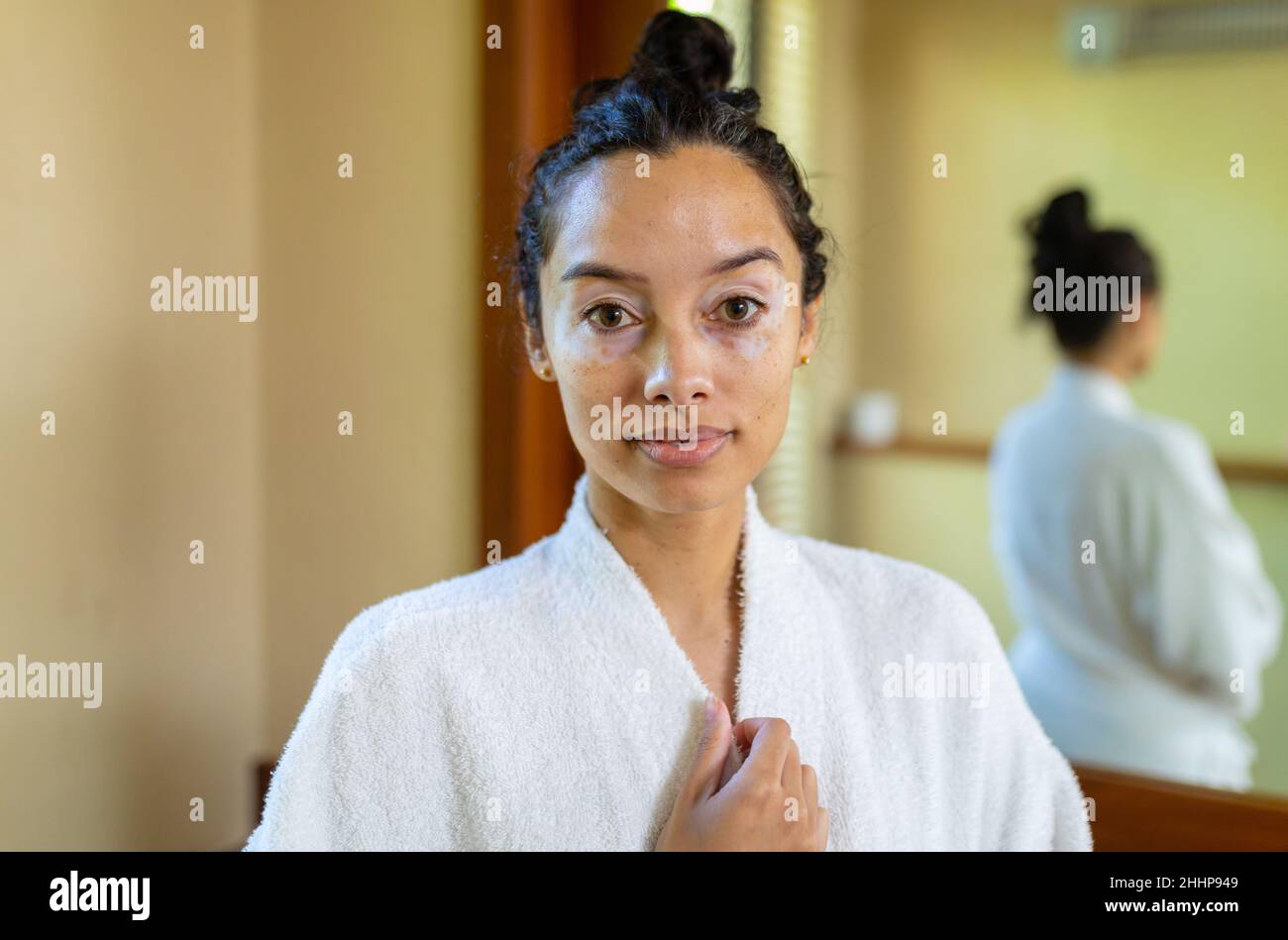 Portrait of biracial young woman wearing bathrobe standing in bathroom. unaltered, routine, lifestyle and vitiligo. Stock Photo