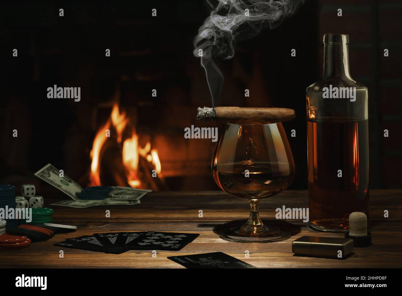 Glass of cognac, cigar, bottle, cards game and chips on the table near the burning fireplace. Rest, pleasure, good luck and dolce vita concept. Stock Photo