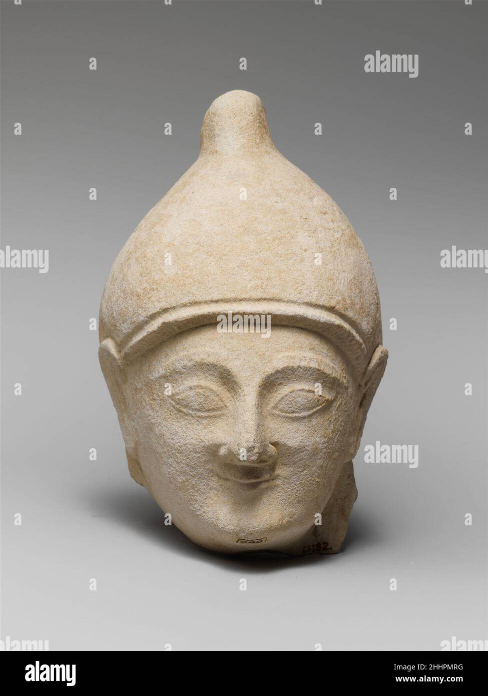 Limestone head of a man wearing a helmet middle or 3rd quarter of the 6th century B.C. Cypriot The face has a smiling expression, the nose is slightly hooked, the eyes almond-shaped, the eyebrows high and thick, the ears schematic, the frontal locks jagged. The tall helmet has a smooth surface and the peak is nothing more than a stump. On the back is just a bit of the hair that projected below the helmet.. Limestone head of a man wearing a helmet  242108 Stock Photo