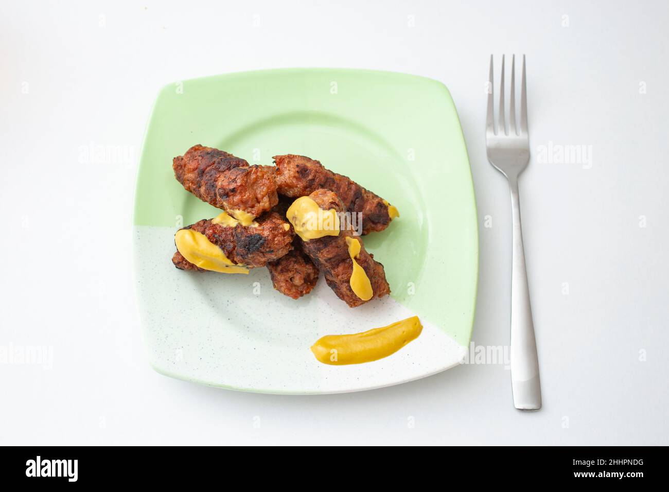 Mici or mitititei (traditional romanian food) with mustard on a ceramic plate, on white background. Hand holding a stainless steel fork Stock Photo