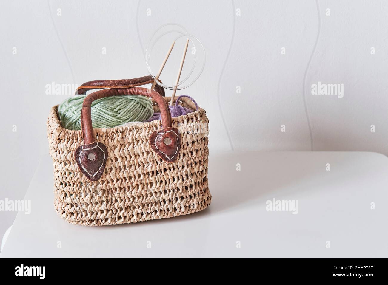 Skeins of yarn and wooden circular knitting needles in handmade straw basket on white table. Stock Photo