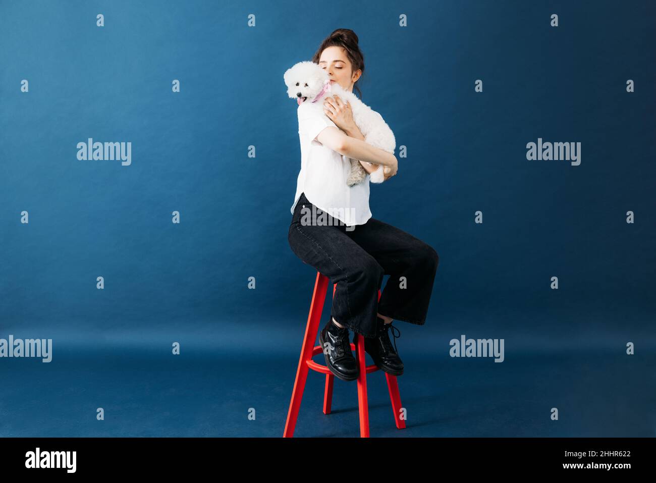 Young woman holding her little dog on hands with closed eyes while sitting on a chair in studio Stock Photo