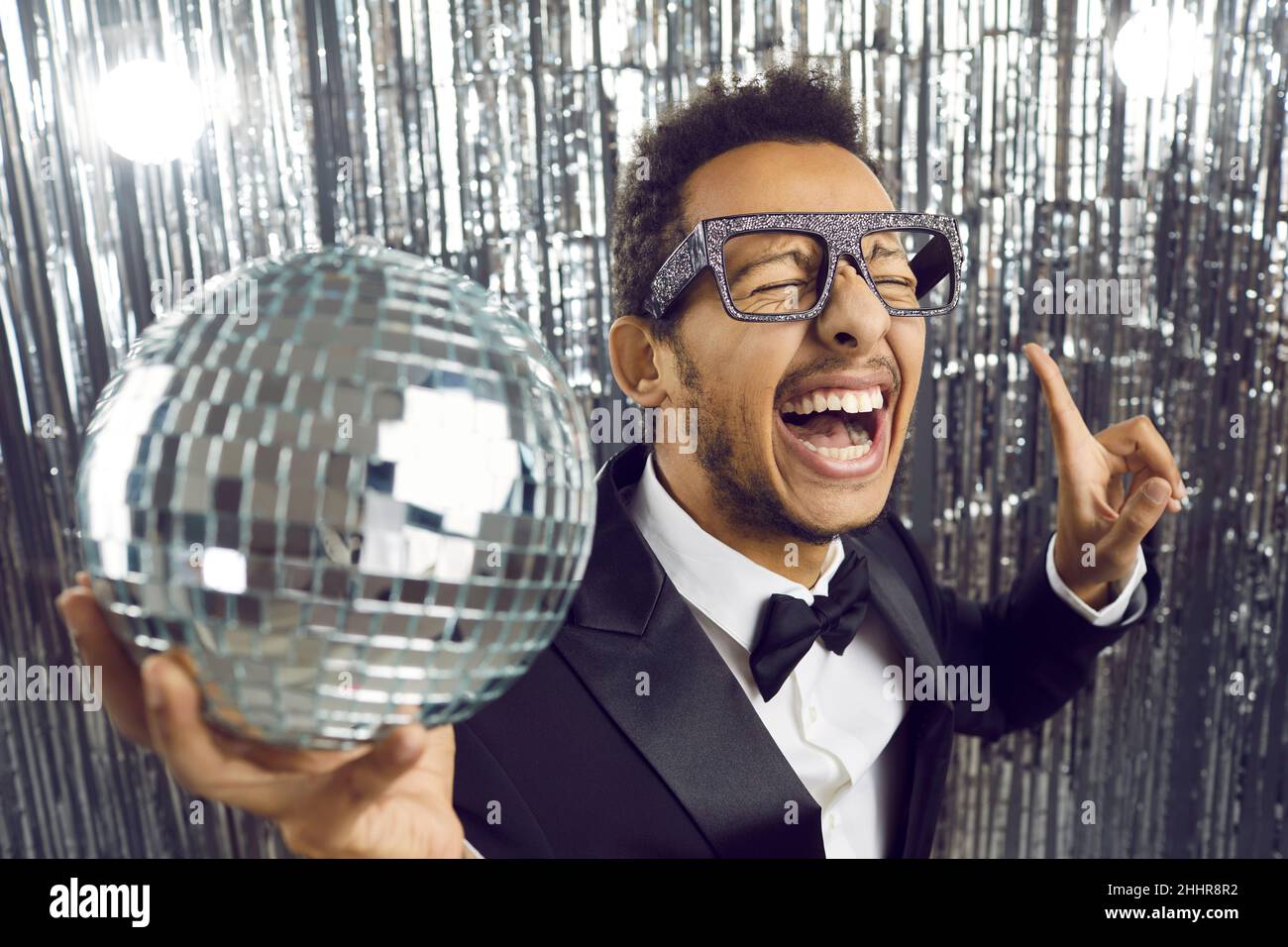 Happy funny ethnic young man in tuxedo and glasses dancing with disco ball at party Stock Photo