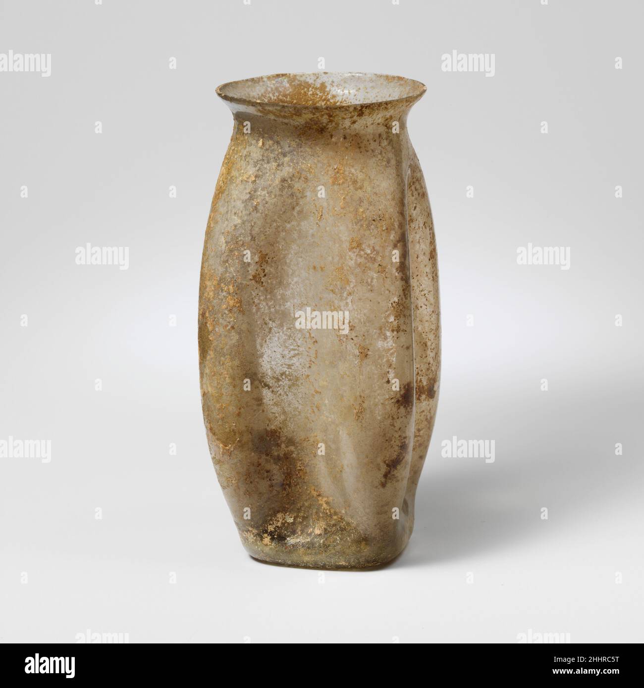 Glass beaker 2nd–3rd century A.D. Roman ColorlessKnocked-off, uneven, flaring rim; concave neck; four-sided body with rounded, convex corners and slightly bulging pad at base; concave bottom.Four deep vertical indents in sides.Intact; many pinprick bubbles; dulling, pitting, patches of iridescent weathering and soil encrustation.. Glass beaker  239856 Stock Photo