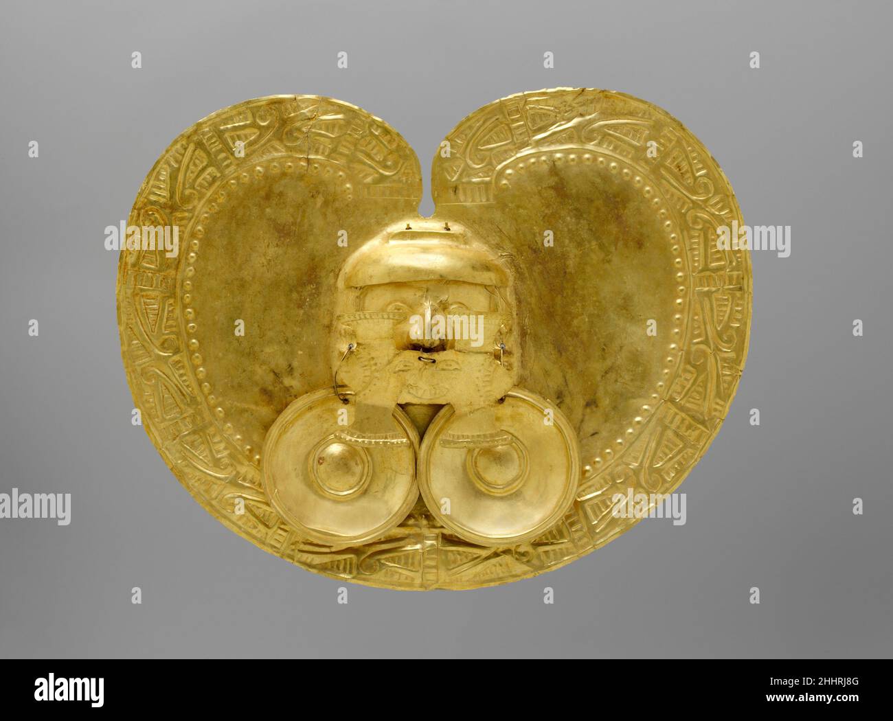 Pectoral with Face 1st–7th century Calima (Yotoco) A large H-shaped nose pendant almost entirely obscures the face at the center of this gold, kidney-shaped pectoral. Both the shape of the pectoral and the form of the nose pendant are hallmarks of Yotoco-period Calima art. The Yotoco period was the second of three societies—Ilama, Yotoco-Malagana, and Sonso—to successively occupy the Cauca Valley region in west-central Colombia. The three societies are known collectively as the Calima culture. The upper portions of the eyes are just visible above the H-shaped pendant, and the head itself is su Stock Photo