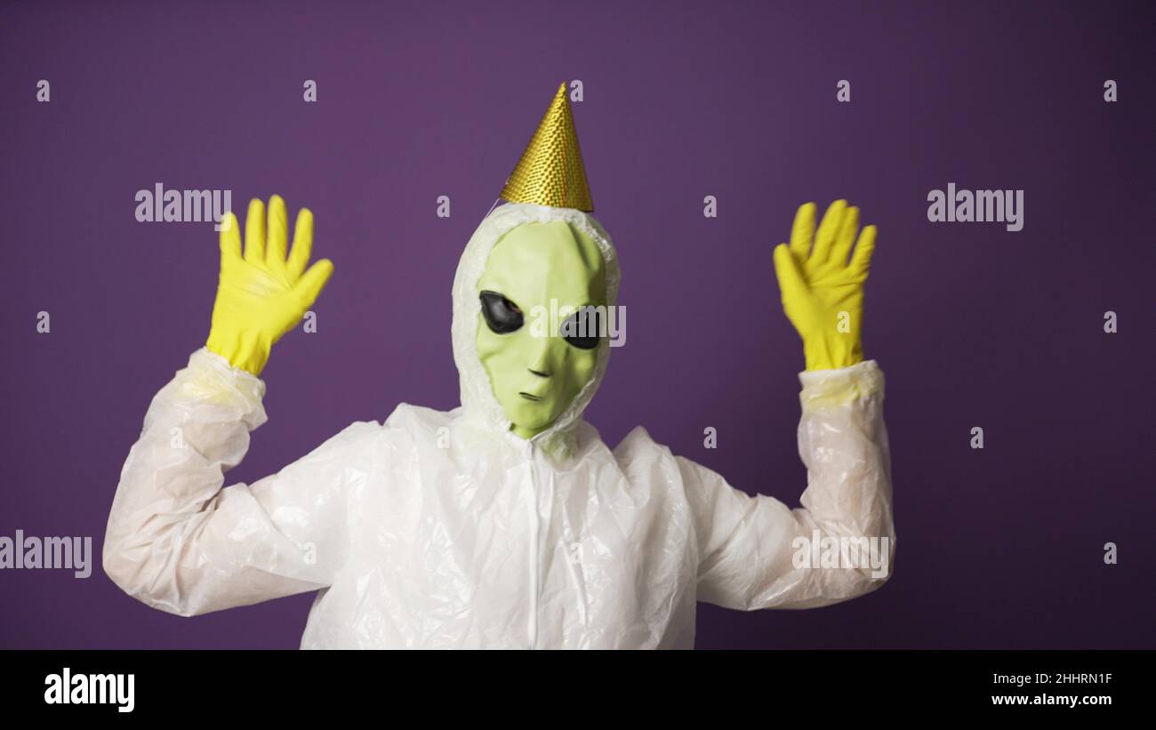 Bright-green alien with big black eye sockets wearing protection costume and gloves with birthday cap dances against violet wall closeup Stock Photo