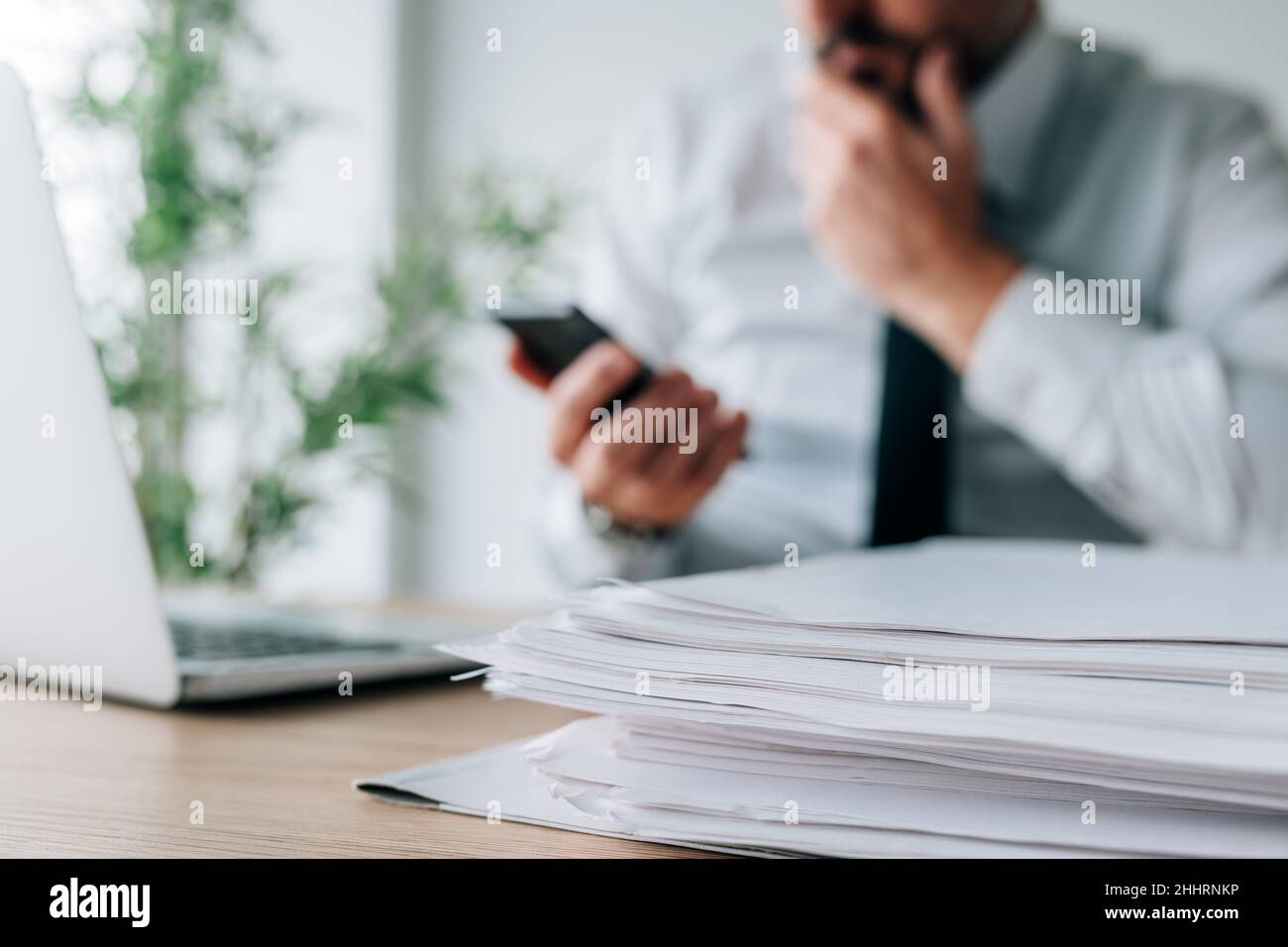 Businessman reading sms text message on smartphone with paperwork pile on office desk, selective focus Stock Photo