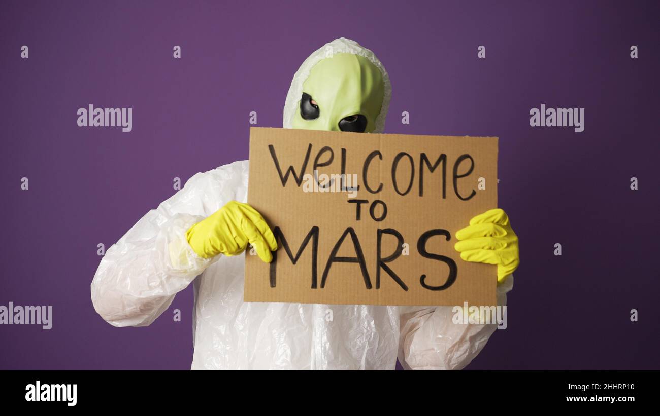 Green alien in protective sanitary costume and yellow gloves welcomes people to planet Mars holding cardboard sign against purple wall closeup Stock Photo