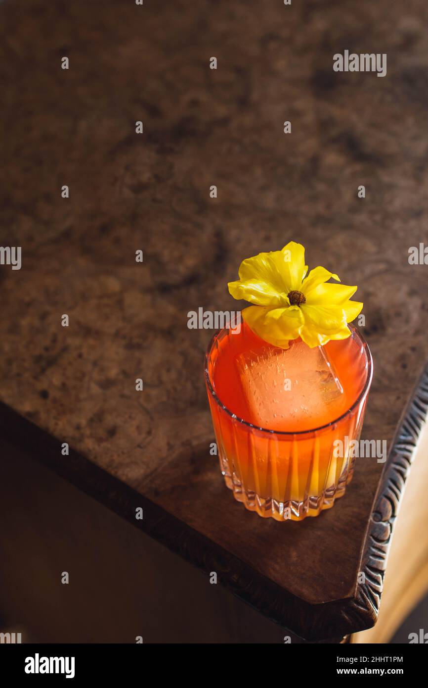 red orange cocktail in crystal rocks glass with yellow flower garnish on burl wood table Stock Photo
