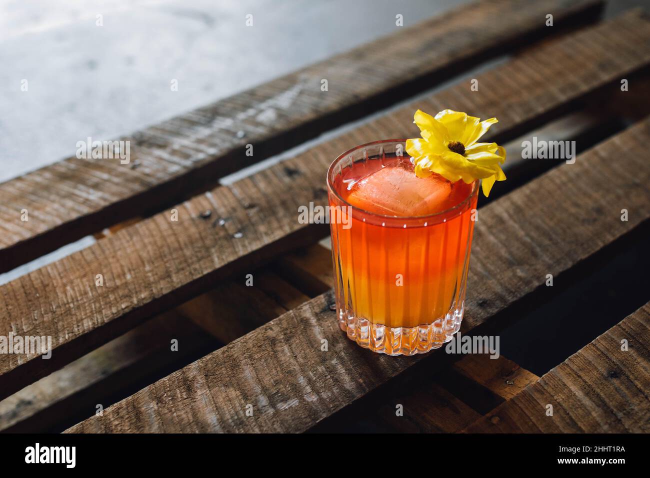 red orange cocktail in crystal rocks glass with yellow flower garnish on wooden pallet table Stock Photo