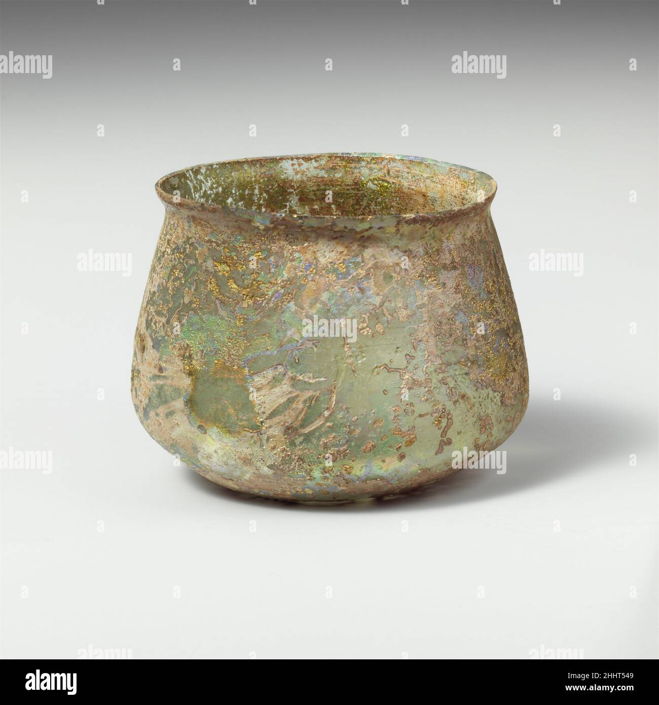 Glass cup 2nd–3rd century A.D. Roman Colorless with blue green tinge.Knocked-off, uneven rim; slightly bulging collar below rim; sides expanding downward, then angled in to join bottom with pushed-in center.Band of faint wheel-abraded horizontal lines on body above angle.Intact; many pinprick bubbles; dulling, deep pitting, and brilliant iridescent weathering on exterior; only faint weathering on interior.. Glass cup  244606 Stock Photo