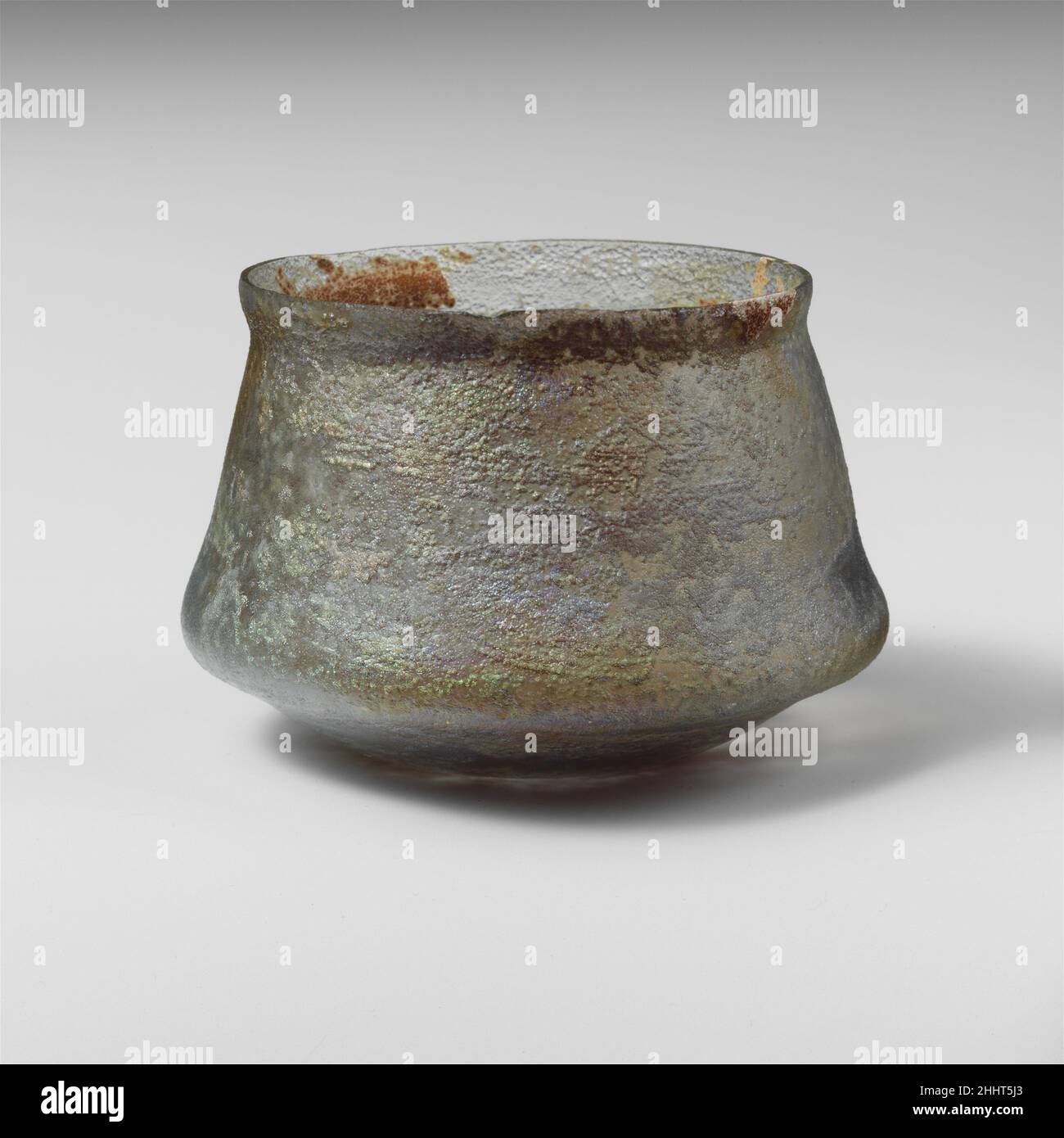 Glass cup 2nd–3rd century A.D. Roman Translucent pale blue green.Knocked-off, uneven rim; slightly bulging collar below rim; straight sides expanding downward, with pronounced projecting ridge below, then angled in to join slightly convex bottom.No trace of any wheel-cut decoration.Intact; many pinprick bubbles; deep pitting, dulling, and iridescence on exterior; creamy brown weathering on interior.. Glass cup  244605 Stock Photo