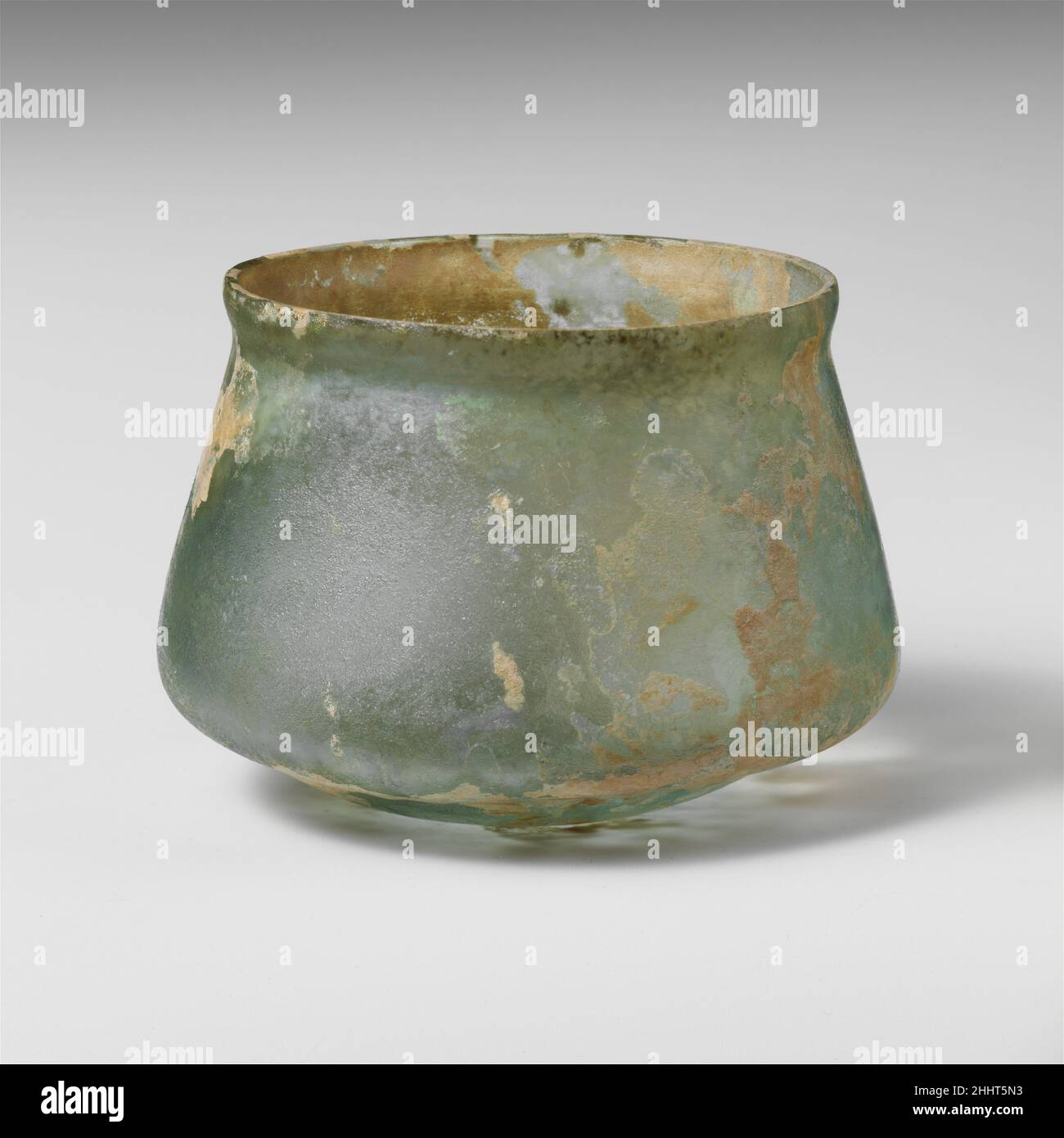 Glass cup 2nd–3rd century A.D. Roman Translucent pale blue green.Knocked-off, slightly uneven rim; slightly bulging collar below rim; sides expanding downward, then angled in to join bottom with pushed-in center.Intact; few bubbles; dulling, faint pitting, iridescence, and patches of creamy weathering.. Glass cup  244608 Stock Photo