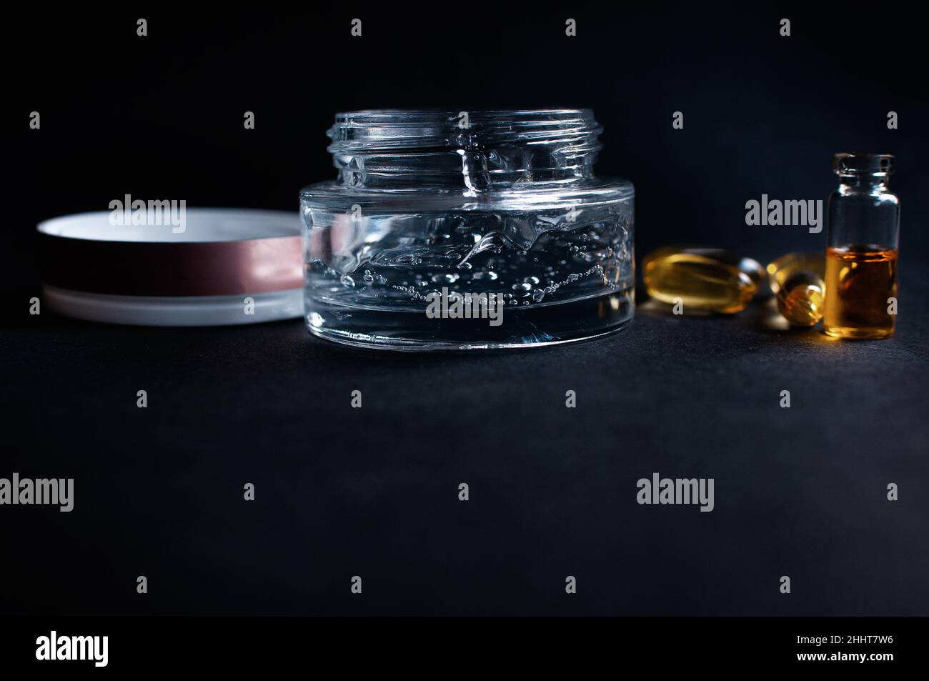 Transparent jar with cosmetic moisturizing gel with hyaluronic acid with bubbles and vitamins in capsules on a black background Stock Photo