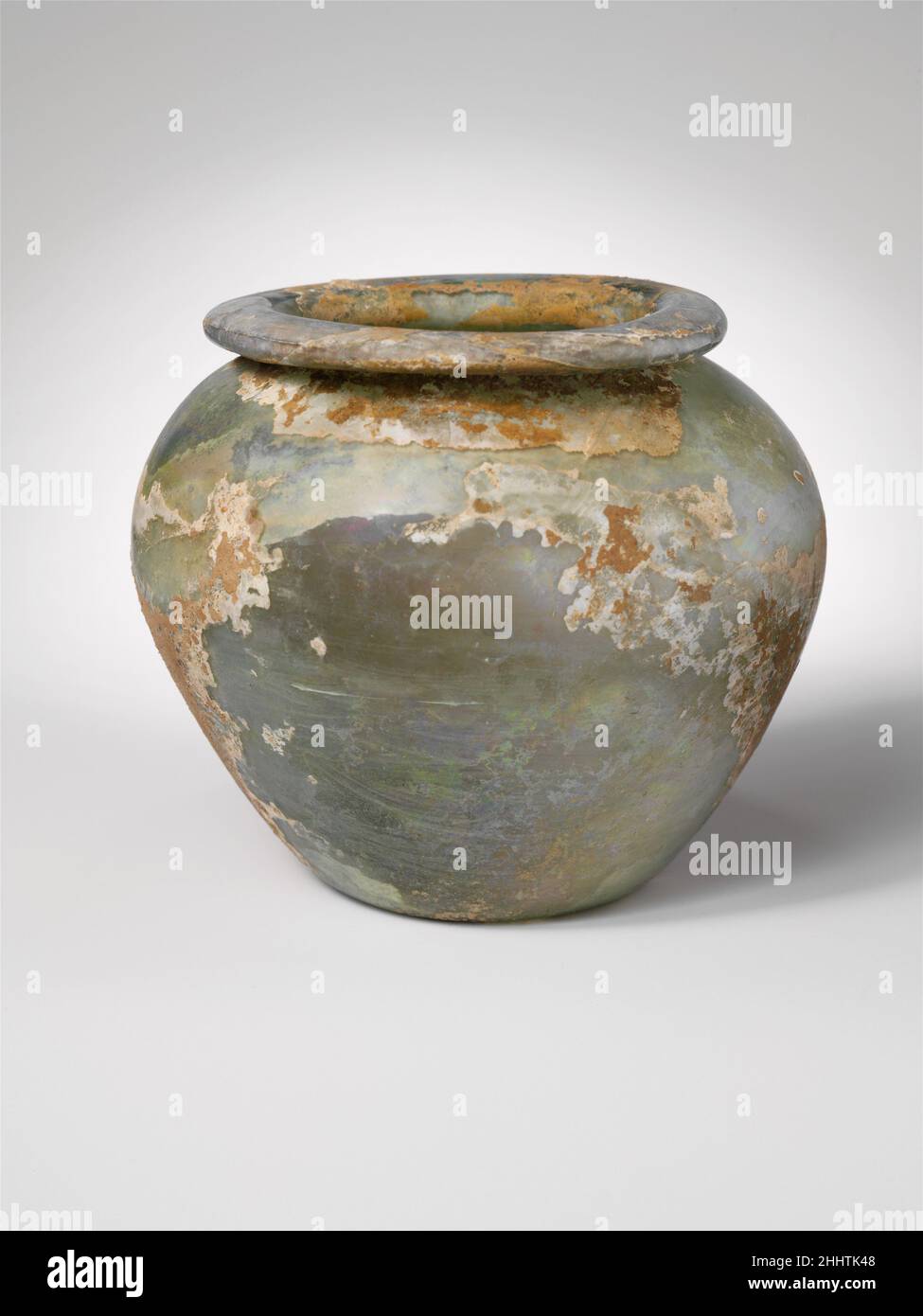 Glass jar mid-1st–2nd century A.D. Roman Colorless with pale blue green tinge.Uneven horizontal rim, with rounded tubular edge, folded out, down, and in, forming collar around neck; ovoid body; concave bottom.Intact, but internal cracks around rim; large and pinprick bubbles, with yellow green impurity streak in rim; some dulling, slight pitting and iridescent weathering, but much of exterior and interior covered with thick limy encrustation and creamy brown weathering.. Glass jar  245176 Stock Photo